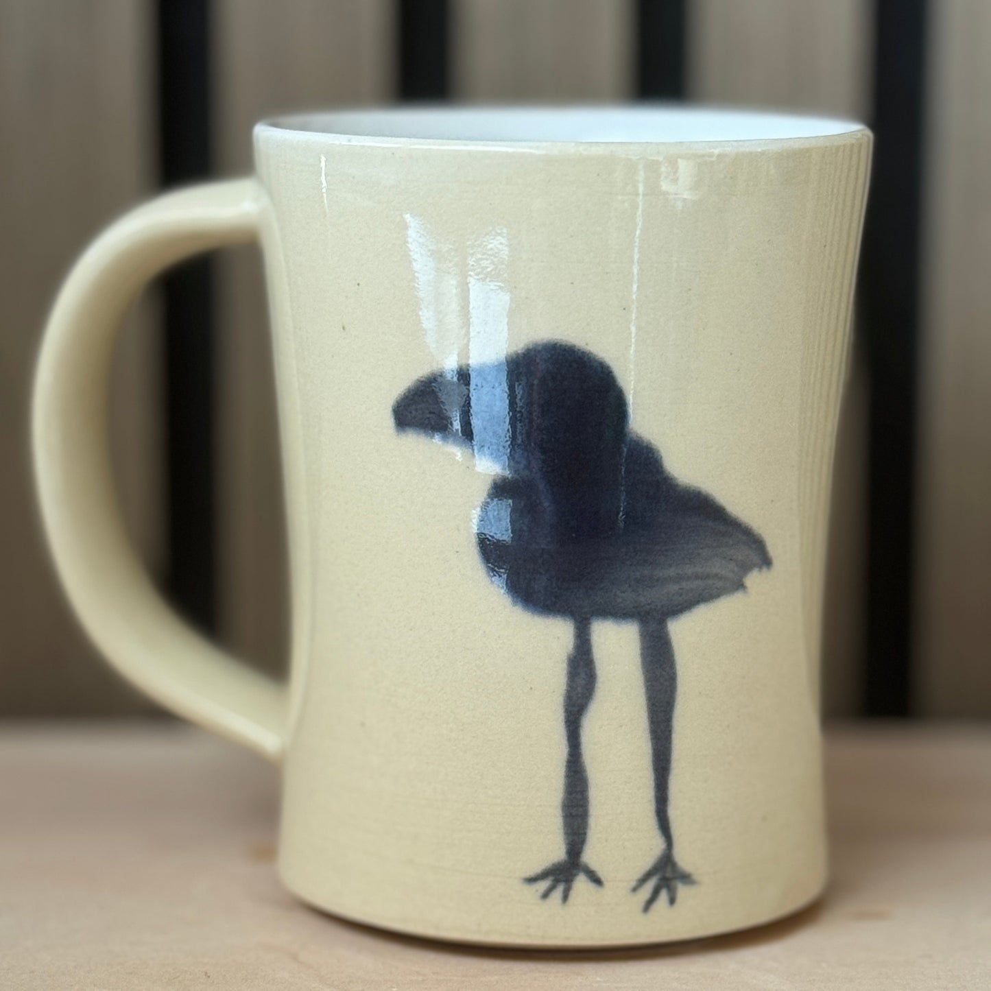 A cream-colored ceramic mug with a black painted bird design, shown on a light wooden surface with a blurred background.