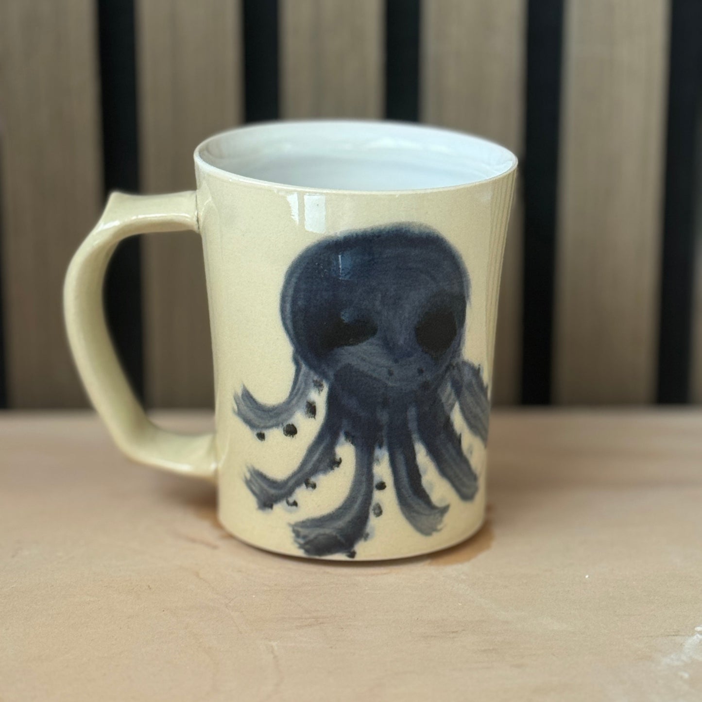 A cream-colored ceramic mug with a dark blue octopus illustration, sitting on a light wooden surface with a wooden slatted background.