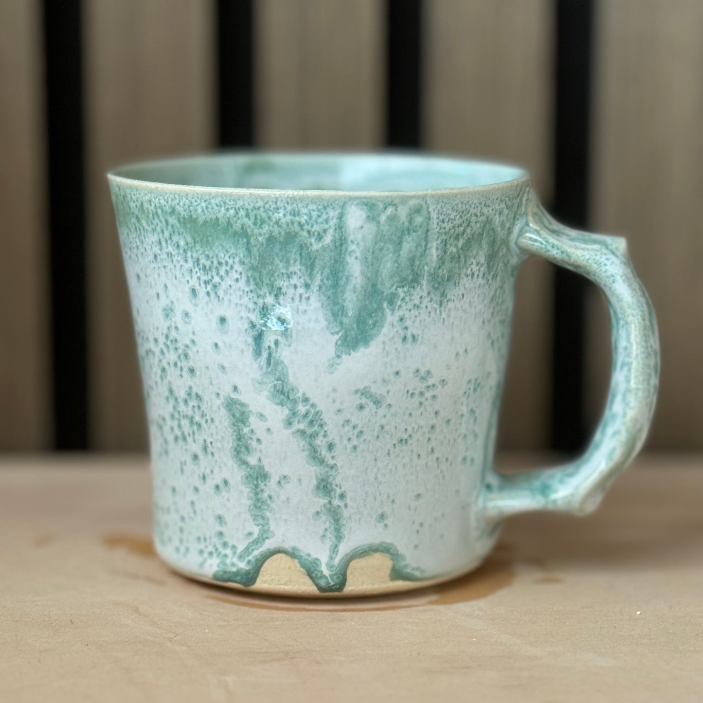 A handmade ceramic mug with a light blue-green and white marbled glaze, sitting on a light wooden surface with a blurred background.