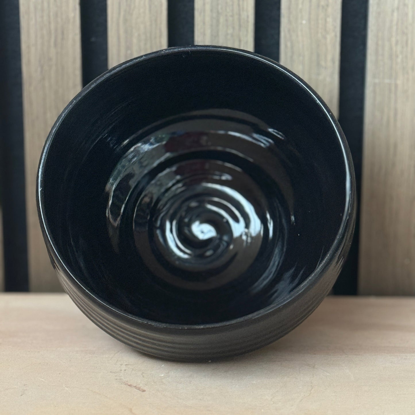 A glossy black ceramic bowl with a ribbed exterior, shown from a top-down perspective on a light wood surface against a black and wood slatted background.