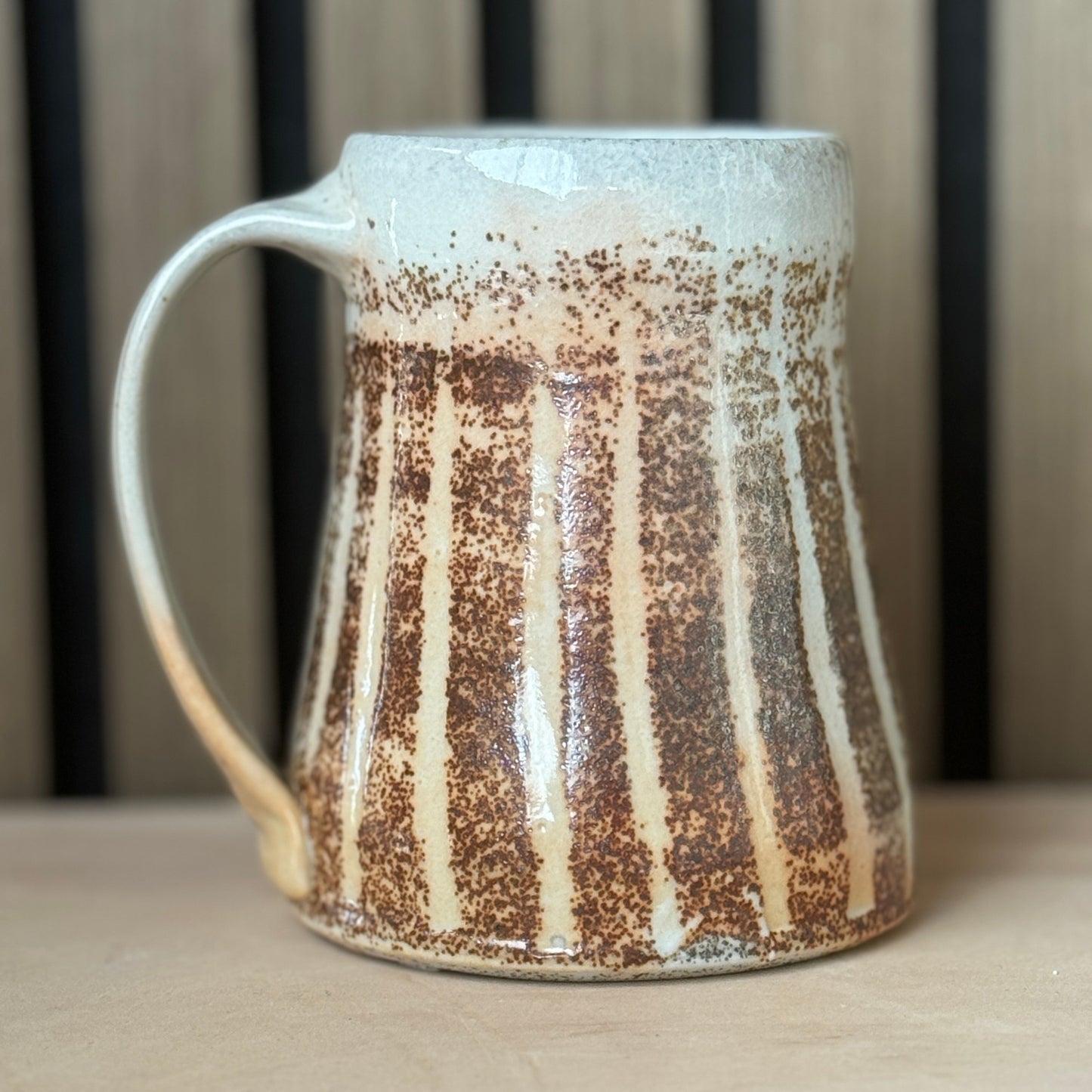 A handmade ceramic mug with a tapered shape, a curved handle, and a textured glaze featuring vertical brown speckled stripes over a cream and light blue-gray base, set on a wooden surface.