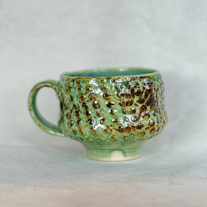 Green ceramic mug with brown speckles on a light gray background