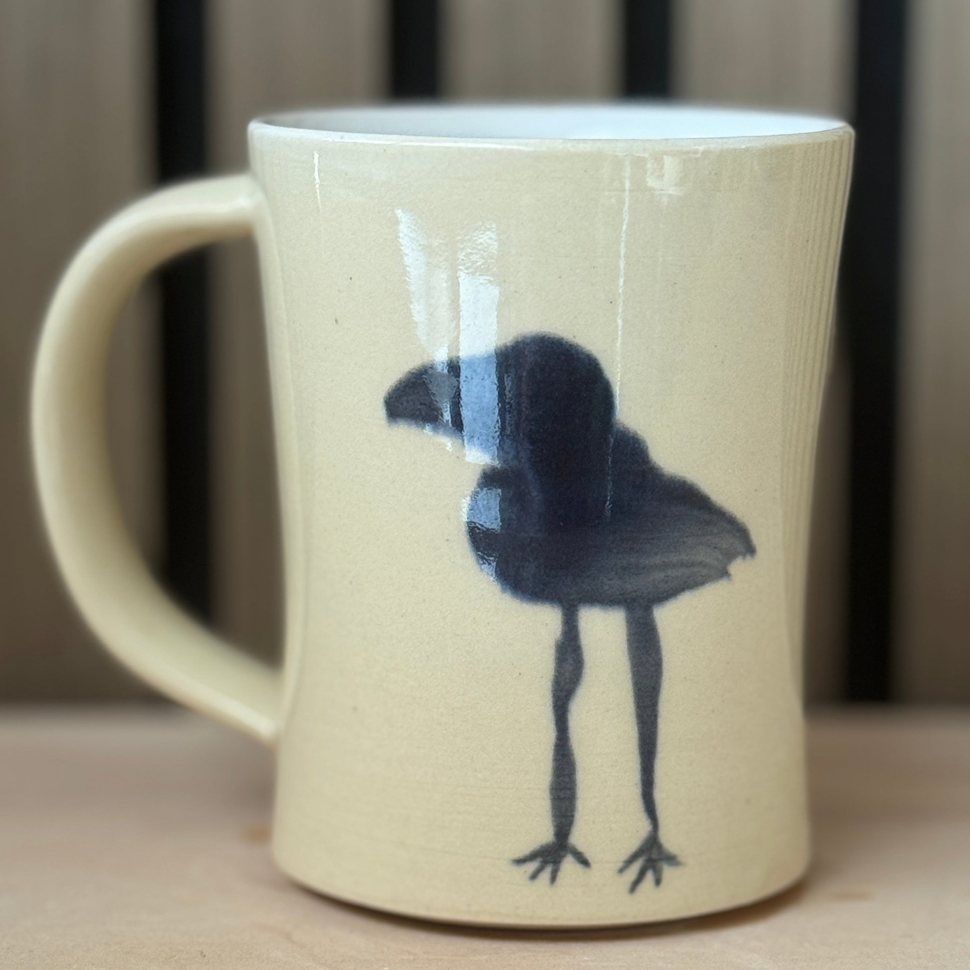A cream-colored ceramic mug with a black painted bird design, shown on a light wooden surface with a blurred background.