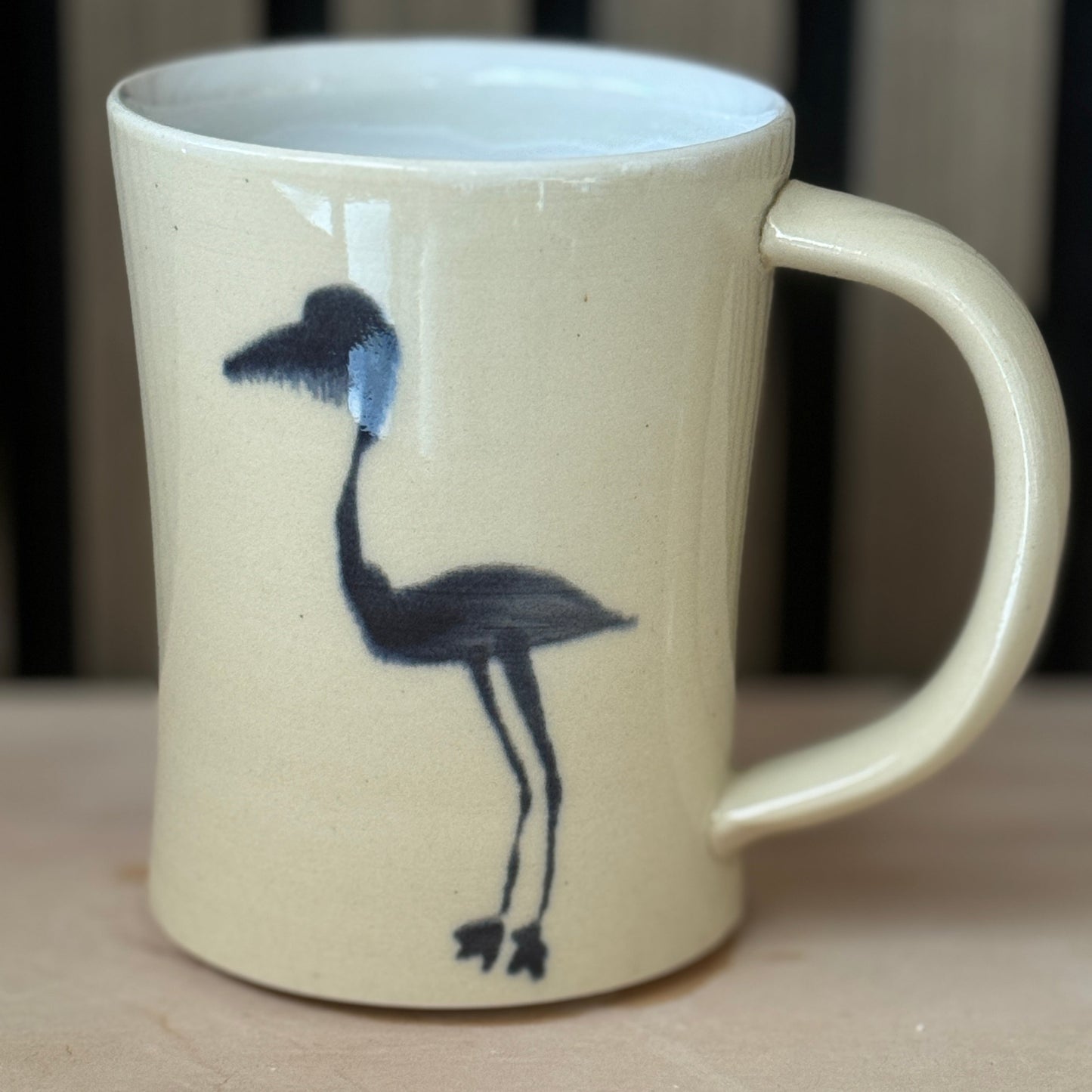A cream-colored ceramic mug with a black bird illustration on the side, set against a dark wood slatted background.