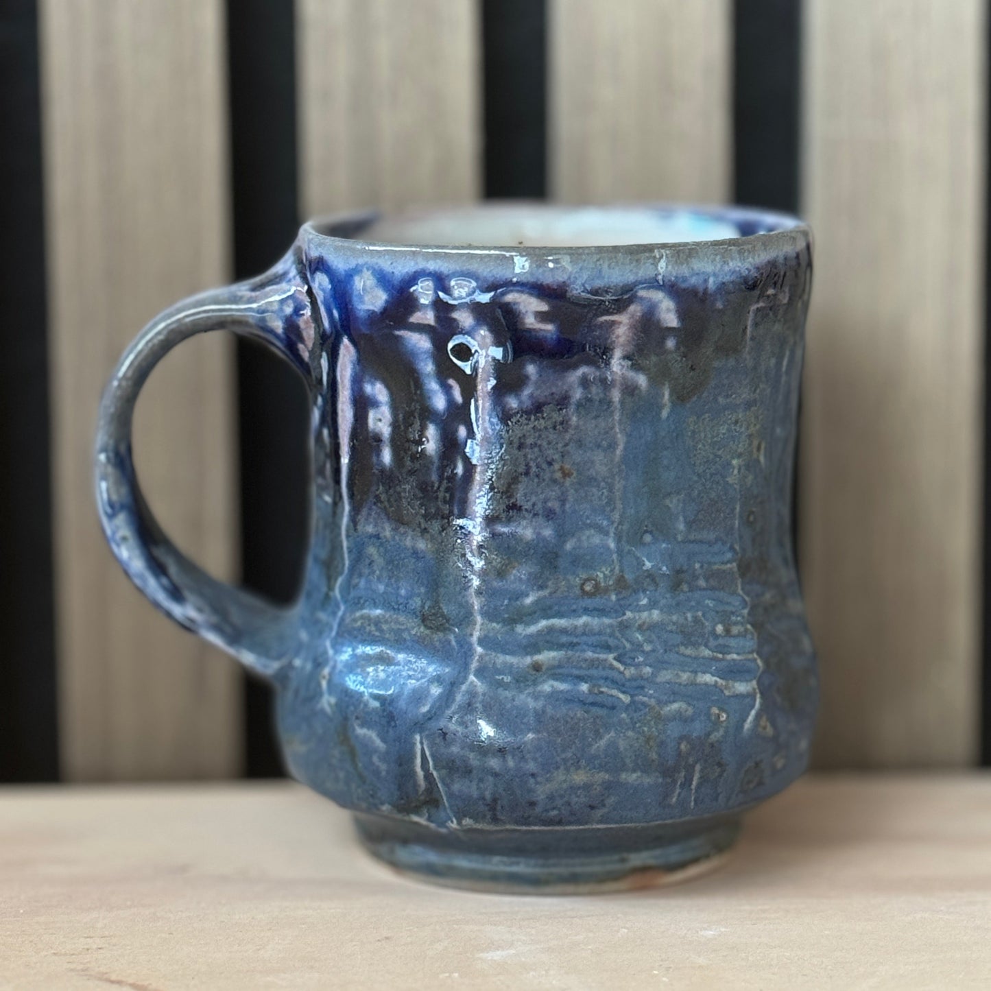 A handmade ceramic mug with a textured, ribbed surface and a dark blue to black glaze with lighter blue highlights, sitting on a light wooden surface against a background of vertical wooden slats.