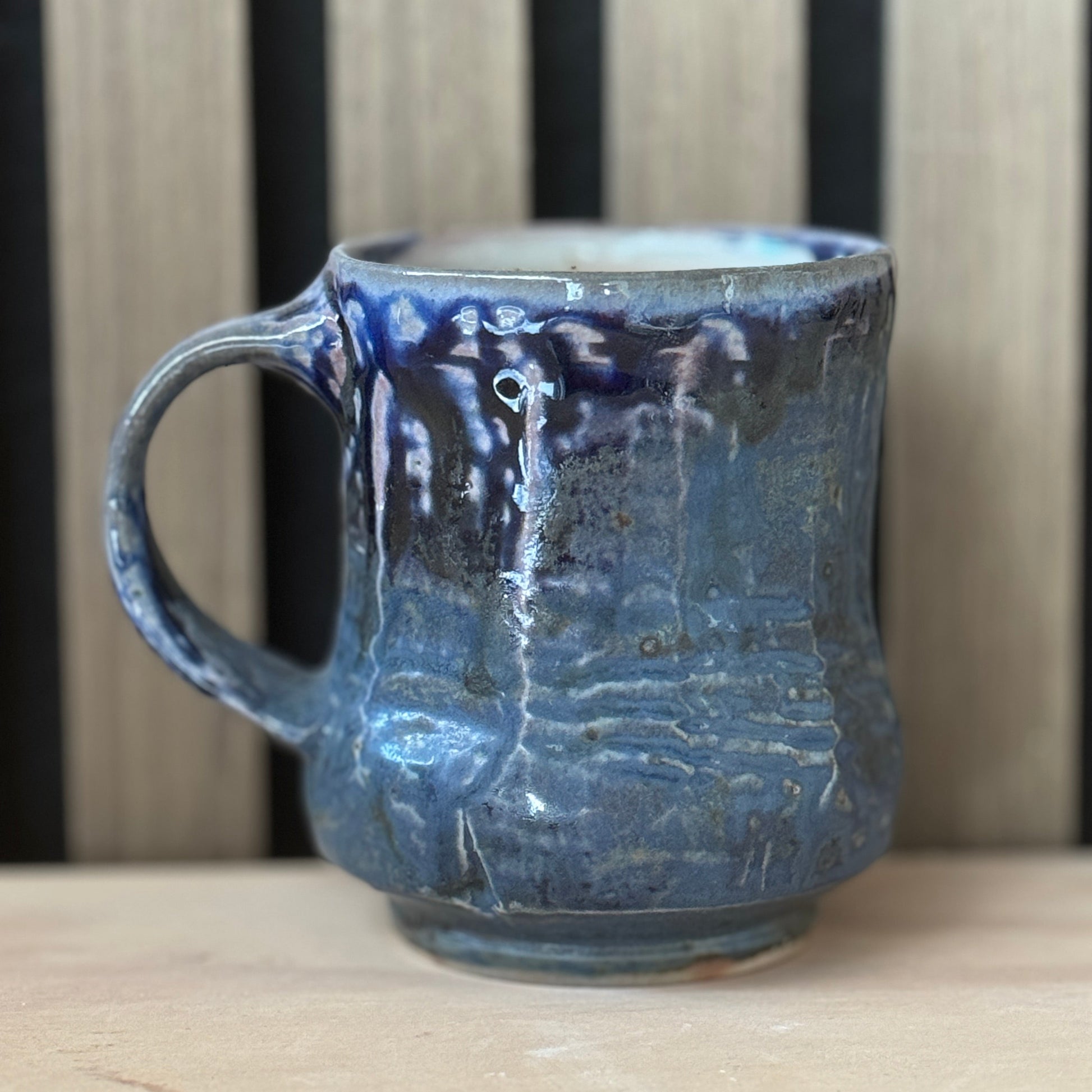 A handmade ceramic mug with a textured, ribbed surface and a dark blue to black glaze with lighter blue highlights, sitting on a light wooden surface against a background of vertical wooden slats.