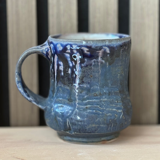 A handmade ceramic mug with a textured, ribbed surface and a dark blue to black glaze with lighter blue highlights, sitting on a light wooden surface against a background of vertical wooden slats.