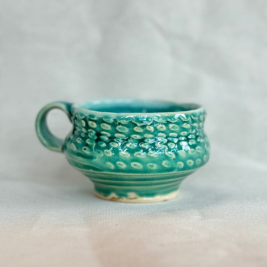 Teal ceramic cup with white speckled pattern on a light gray background