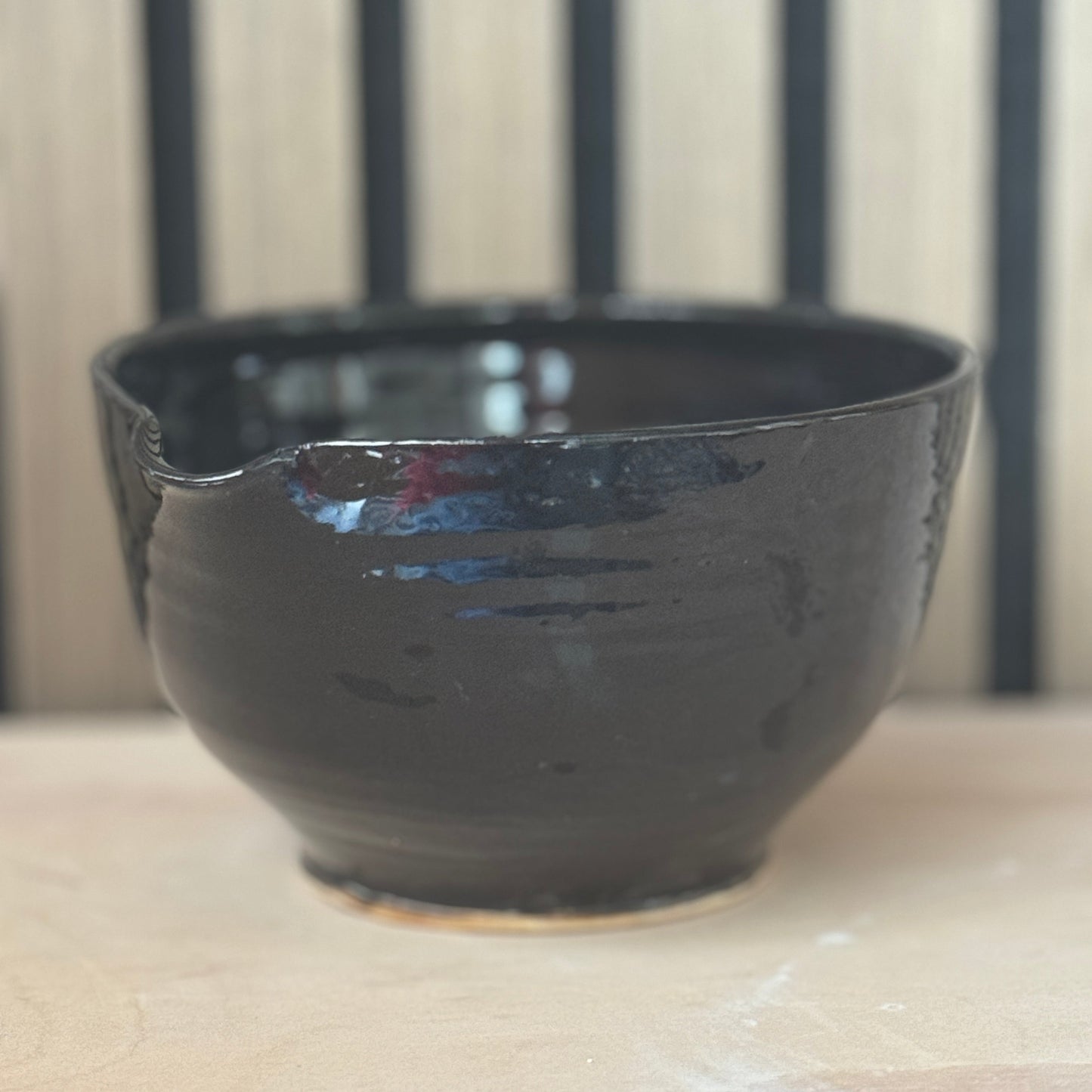 A handmade dark brown ceramic bowl with a glossy finish and a small notch on the rim, resting on a light wooden surface.