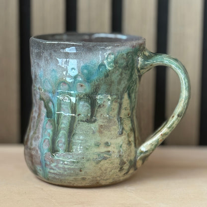 A handmade ceramic mug with a rustic, textured glaze featuring green, teal, and brown drip patterns, set against a background of vertical wooden slats.