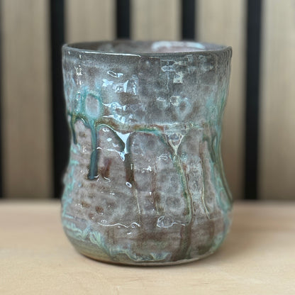 A handmade ceramic cup with a textured, hammered finish and a green glaze drip design, sitting on a light wood surface against a wooden slatted background.
