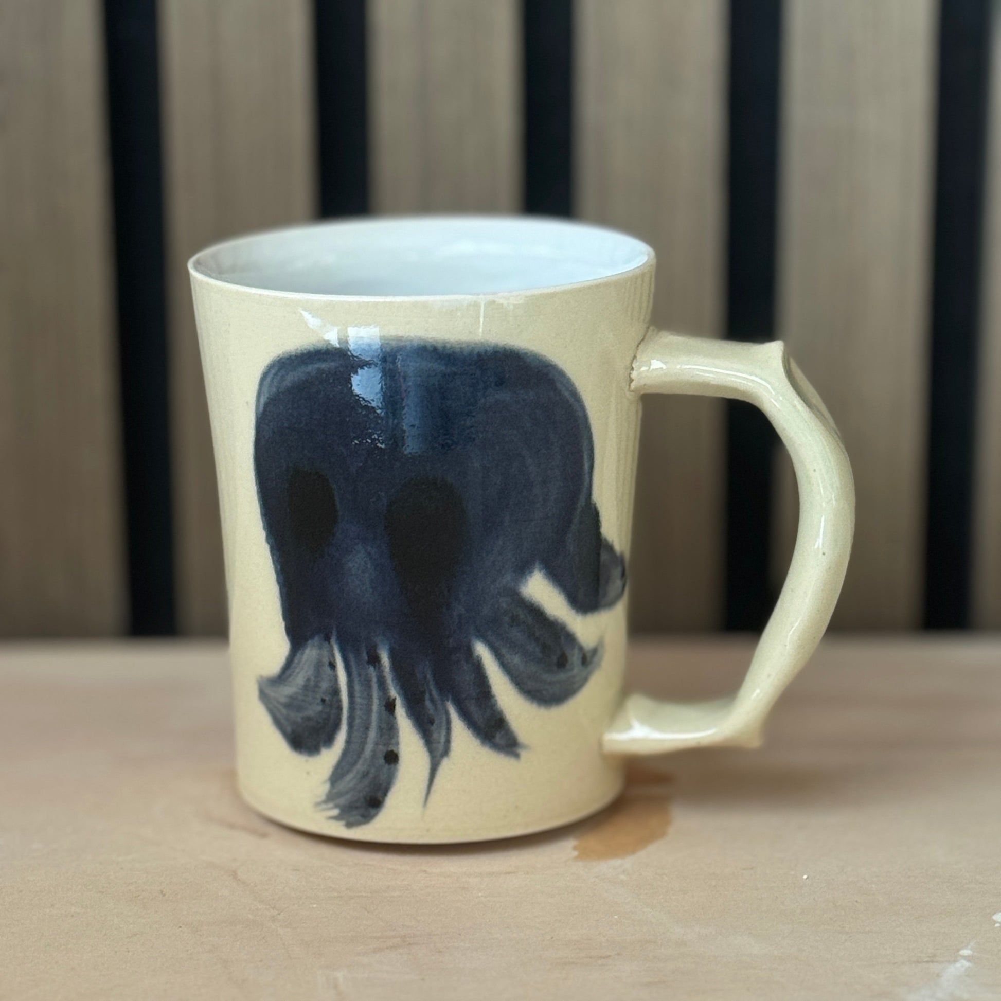 A cream-colored ceramic mug with a dark navy blue skull design, resting on a light wooden surface against a dark wood slatted background.