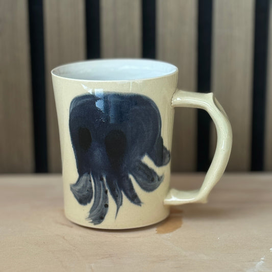 A cream-colored ceramic mug with a dark navy blue skull design, resting on a light wooden surface against a dark wood slatted background.