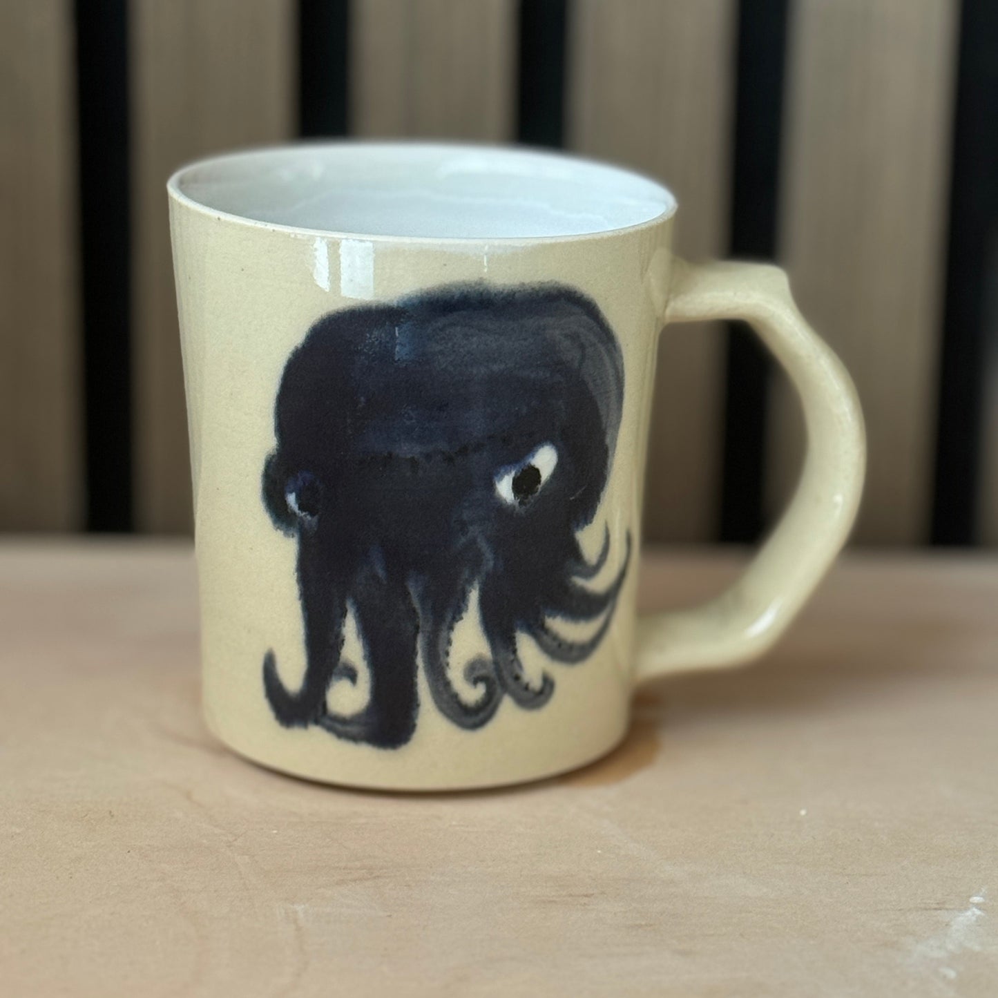 A cream-colored ceramic mug featuring a hand-painted black octopus design, resting on a light wooden surface with a dark slatted background.