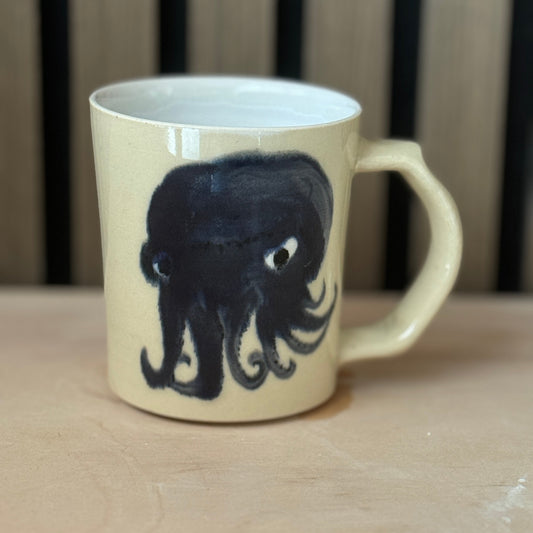 A cream-colored ceramic mug featuring a hand-painted black octopus design, resting on a light wooden surface with a dark slatted background.