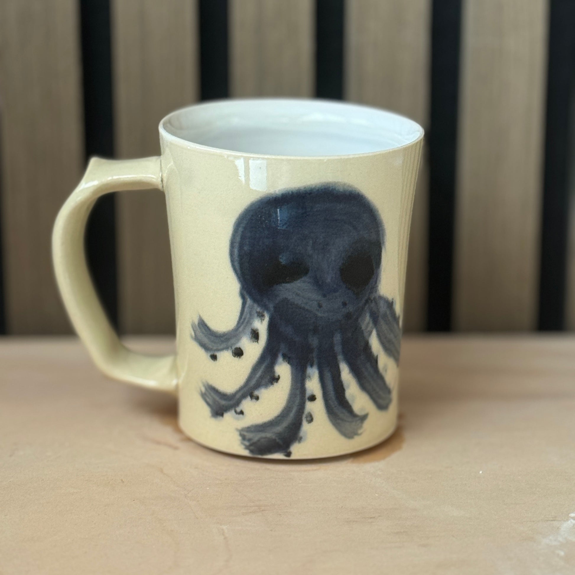 A cream-colored ceramic mug with a dark blue octopus illustration, sitting on a light wooden surface with a wooden slatted background.