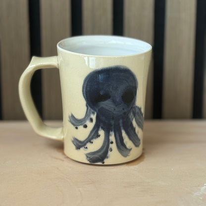 A cream-colored ceramic mug with a dark blue octopus illustration, sitting on a light wooden surface with a wooden slatted background.