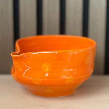 An orange ceramic bowl with a glossy finish and subtle yellow floral patterns, set against a background of vertical dark slats and a light beige surface.