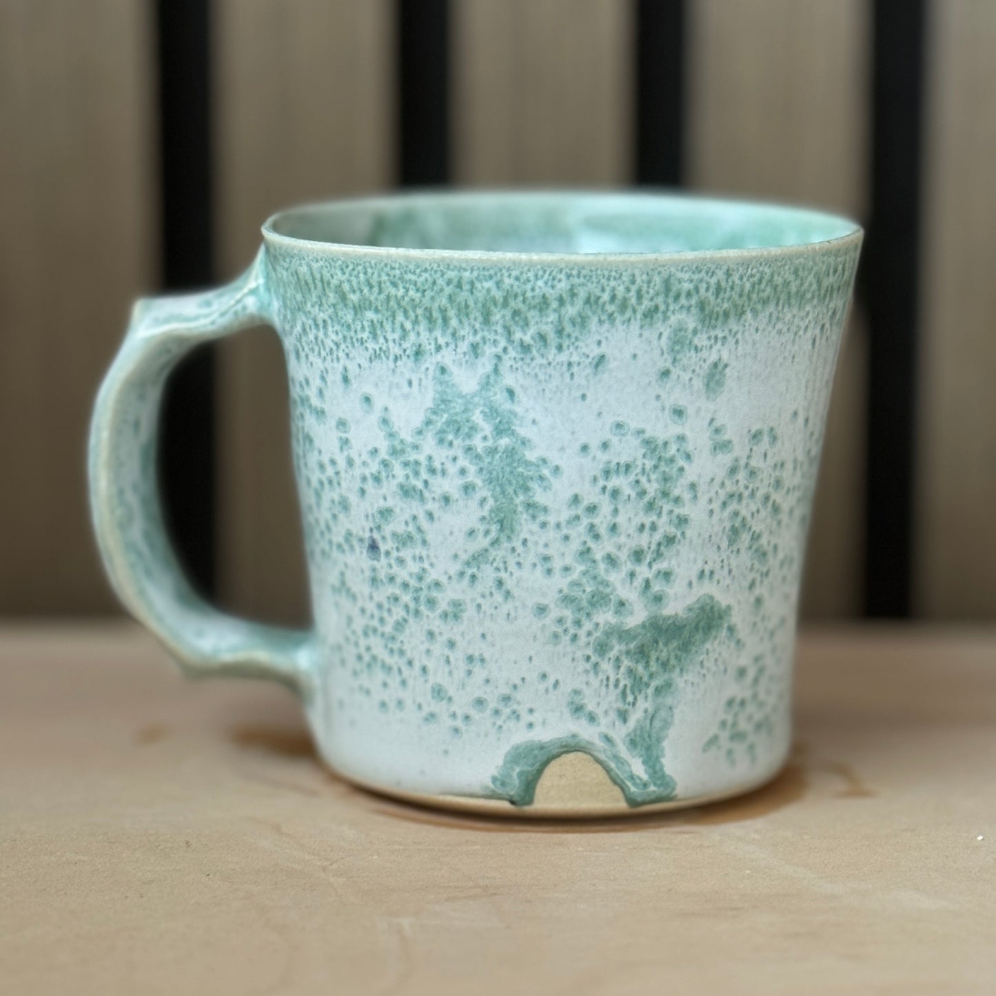 A handmade ceramic mug with a speckled mint green and white glaze, resting on a light wooden surface with a blurred background.