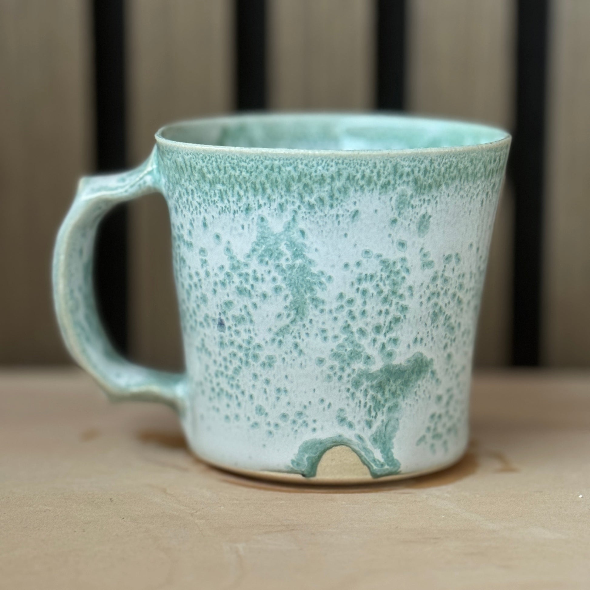 A handmade ceramic mug with a speckled mint green and white glaze, resting on a light wooden surface with a blurred background.