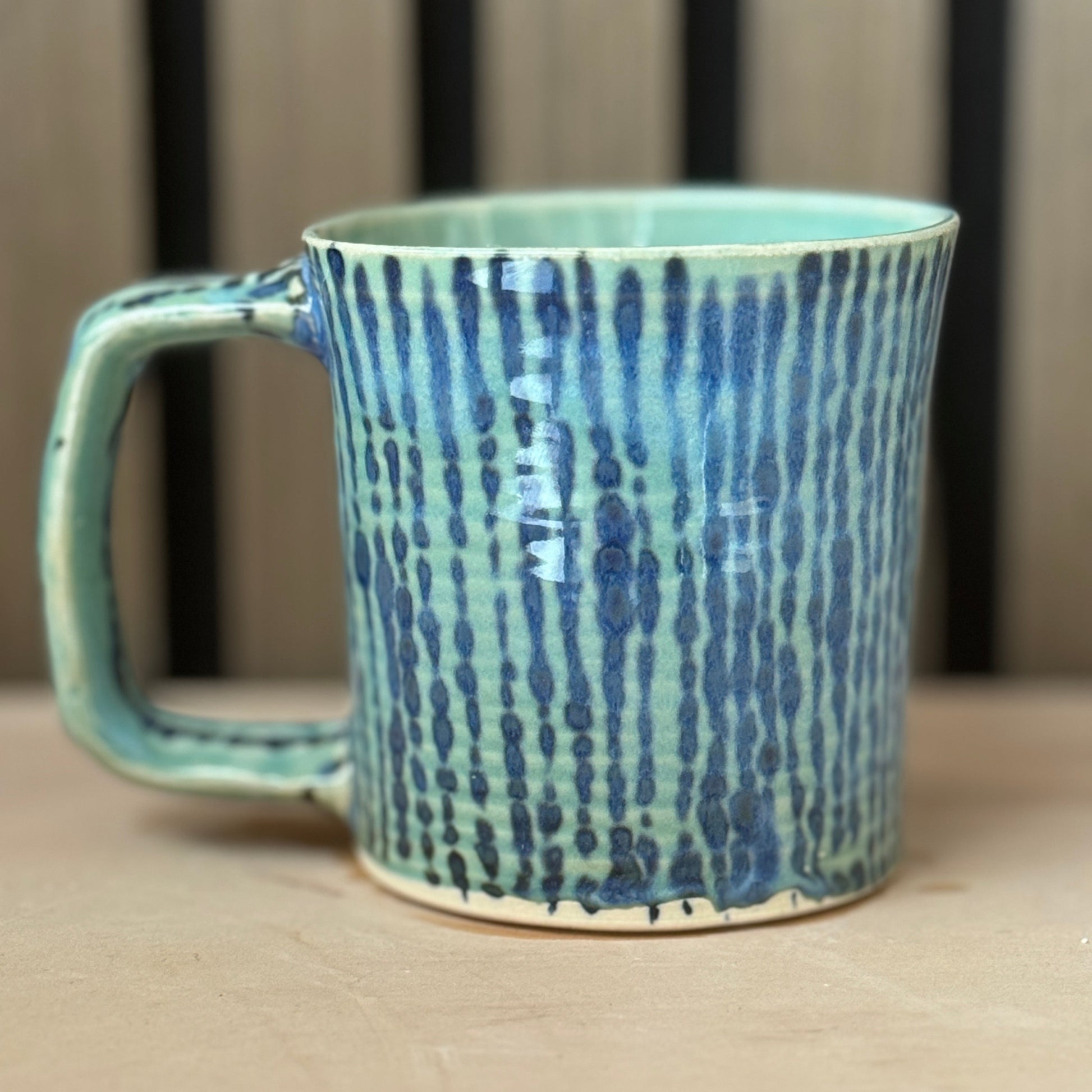 A handmade ceramic mug with a textured vertical blue and green glaze, resting on a light wooden surface with a blurred wooden background.