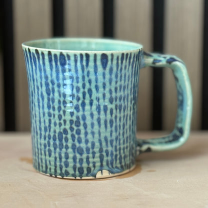 A handmade ceramic mug with a textured vertical dotted pattern in shades of blue and turquoise, resting on a light wooden surface.