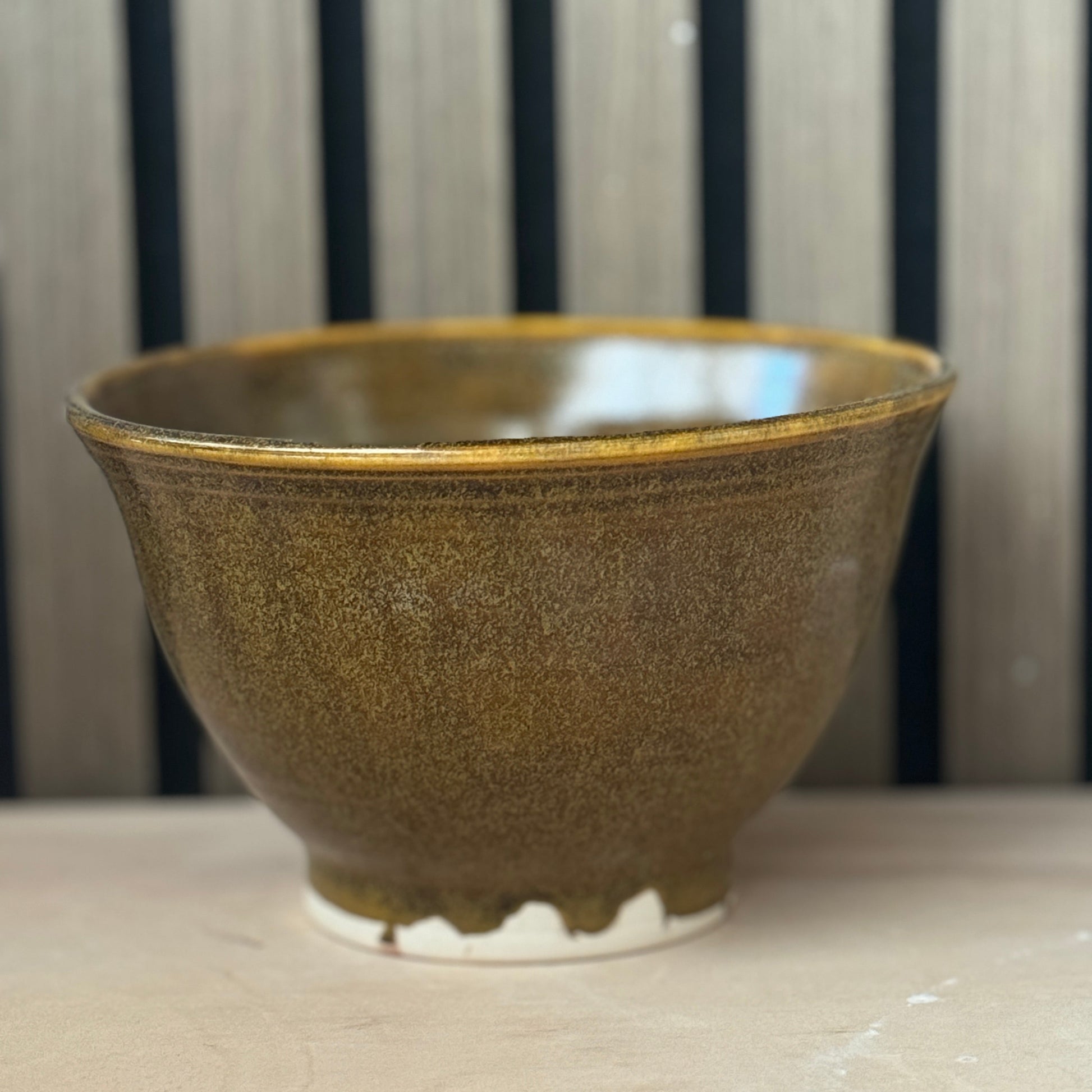 A handmade ceramic bowl with a textured, speckled brown and gold glaze, sitting on a light wooden surface against a background of vertical dark wood slats.