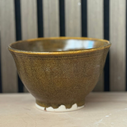 A handmade ceramic bowl with a textured, speckled brown and gold glaze, sitting on a light wooden surface against a background of vertical dark wood slats.
