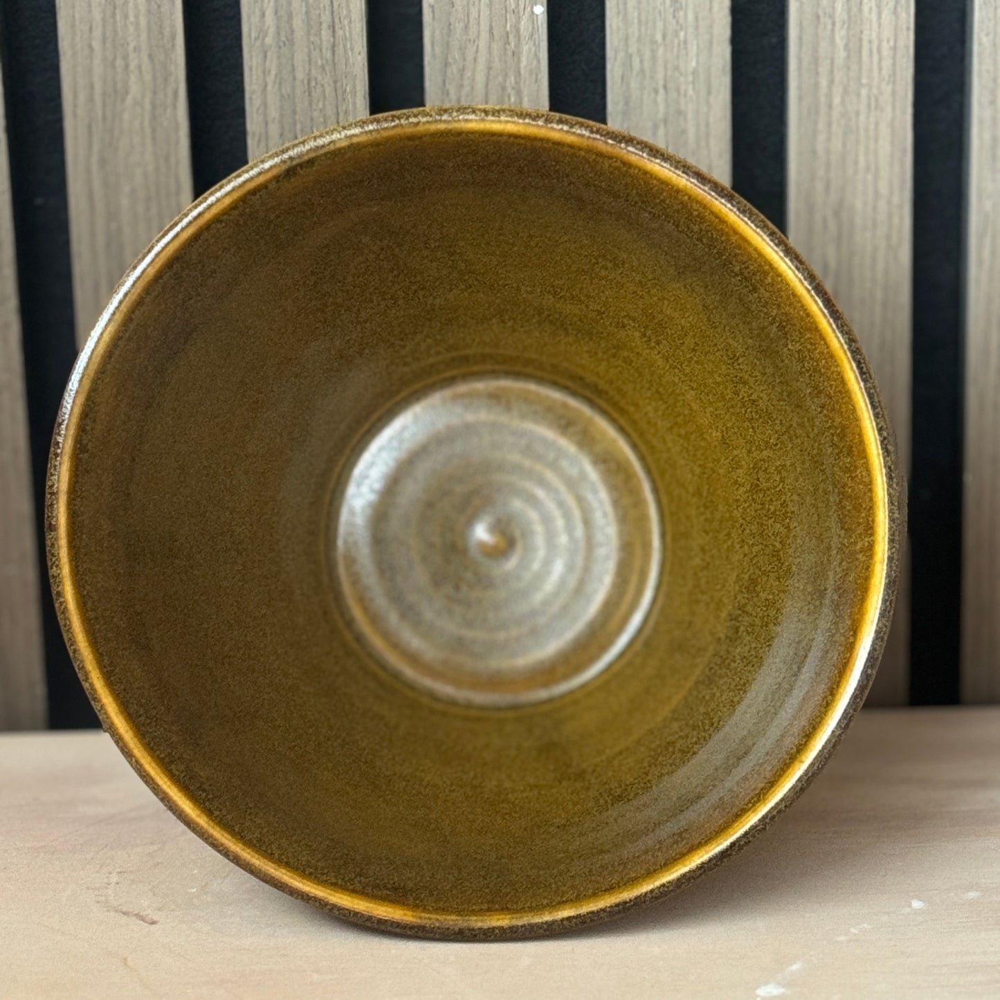 A handmade ceramic bowl with a textured, glossy olive-brown glaze, photographed from a top-down perspective on a light wooden surface against a black and wood slatted background.