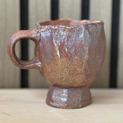 A rustic handmade ceramic mug with a textured brown and reddish-brown glaze, featuring a thick handle and a small pedestal base, set against a blurred background of vertical wooden slats.