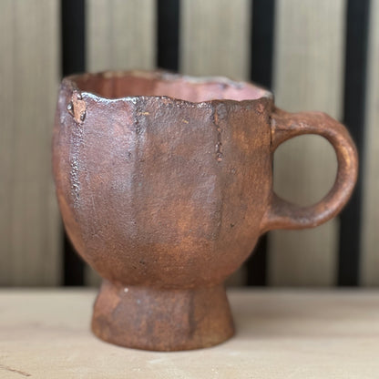 A rustic handmade ceramic mug with a textured terracotta finish, featuring a flared rim, a sturdy base, and a single loop handle, set against a neutral background.