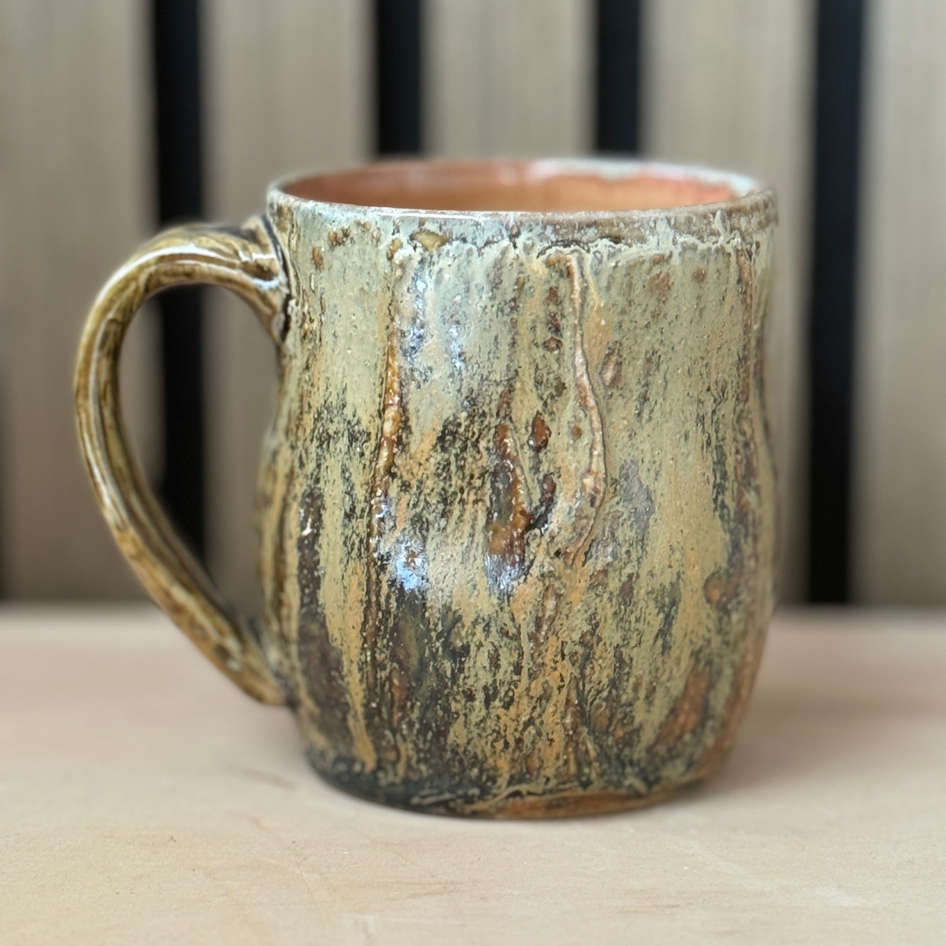 A rustic handmade ceramic mug with a textured, bark-like glaze in earthy brown, beige, and gold tones, set against a neutral background.