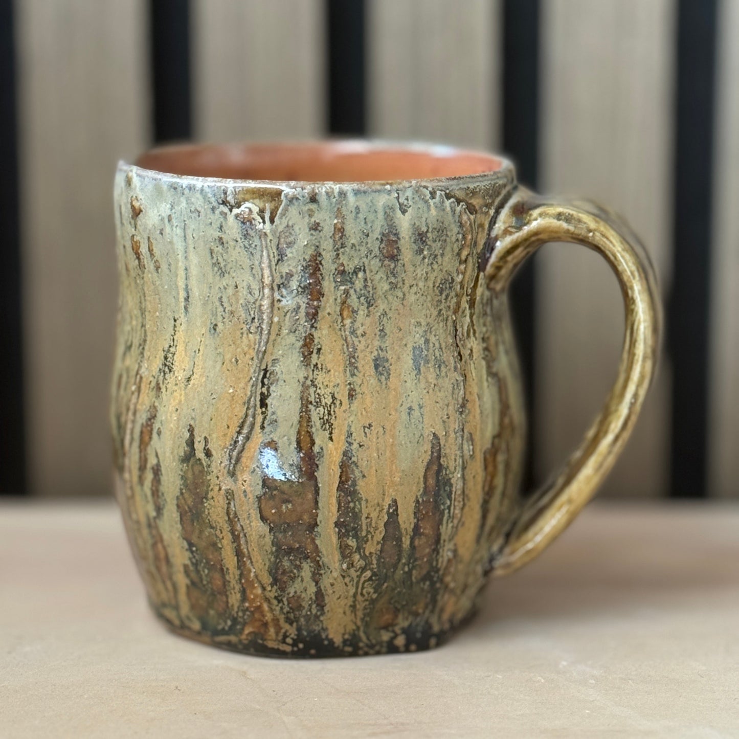 A rustic ceramic mug with a textured, bark-like glaze in earthy brown, beige, and green tones, featuring a warm terracotta-colored interior and a sturdy handle, set against a blurred background of vertical wooden slats.