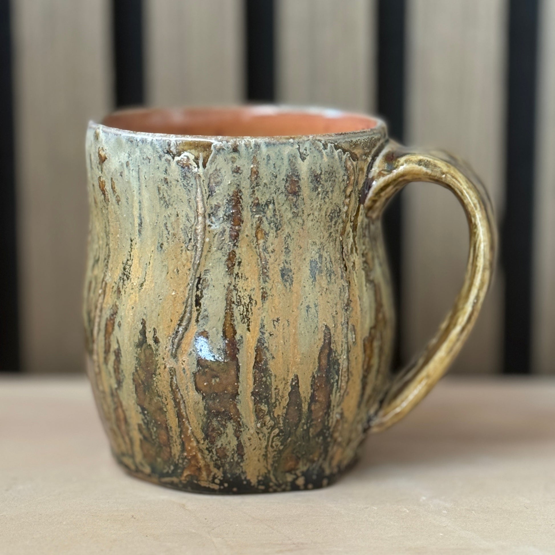 A rustic ceramic mug with a textured, bark-like glaze in earthy brown, beige, and green tones, featuring a warm terracotta-colored interior and a sturdy handle, set against a blurred background of vertical wooden slats.