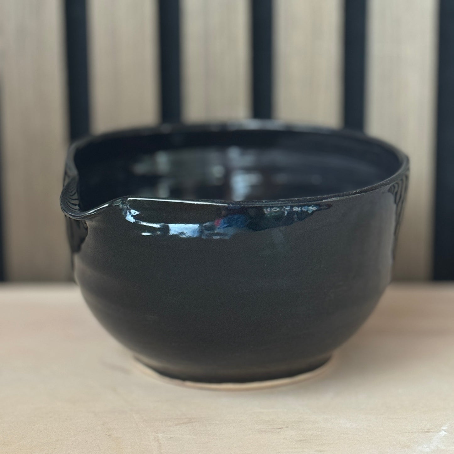 A glossy black ceramic bowl with a small pouring spout, resting on a light wooden surface against a background of vertical dark wood slats.