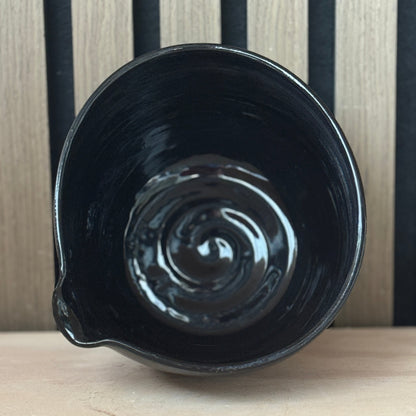 A glossy black ceramic bowl with a small pouring spout, shown from a top-down perspective on a light wooden surface against a black and wood slatted background.