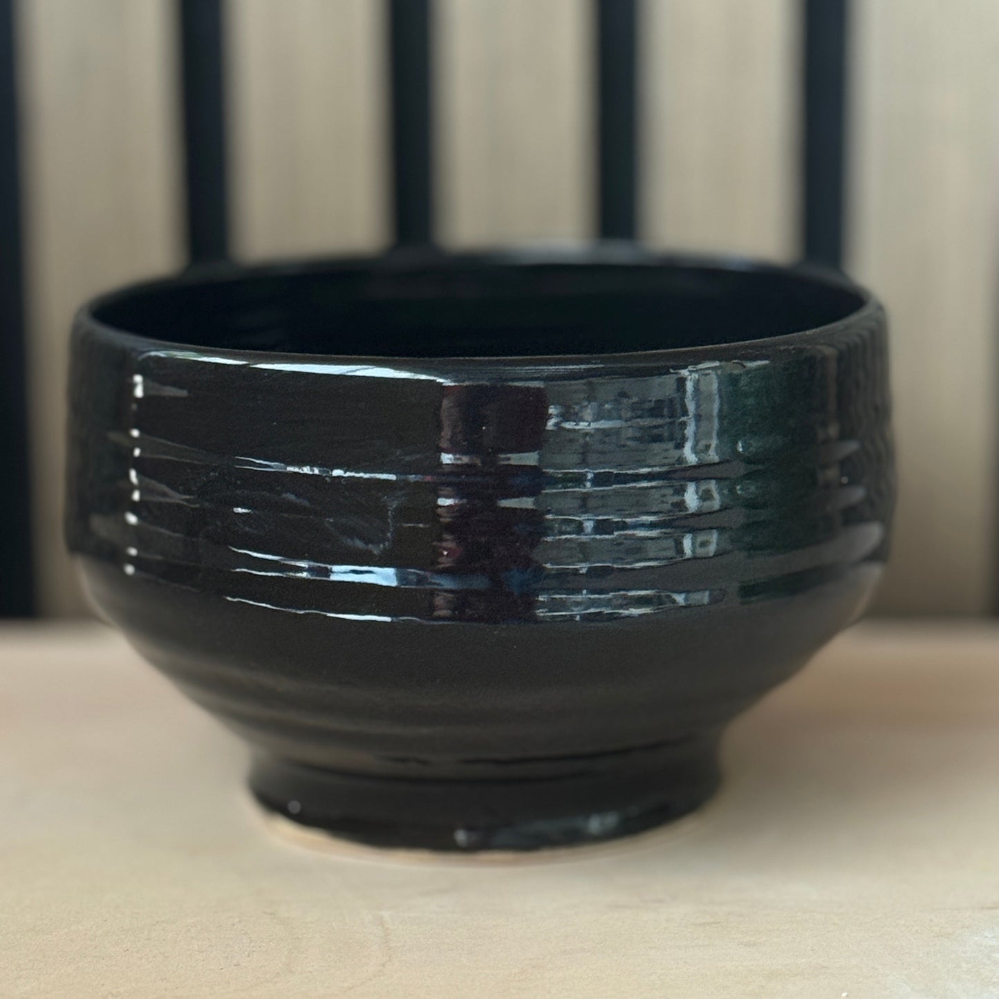 A glossy black ceramic bowl with horizontal textured ridges, shown on a light beige surface with a blurred background of vertical dark lines.