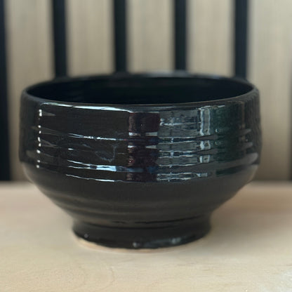 A glossy black ceramic bowl with horizontal textured ridges, shown on a light beige surface with a blurred background of vertical dark lines.