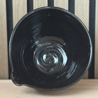 A glossy black ceramic bowl with a small pouring spout, photographed from a top-down angle against a background of vertical black and light wood slats.