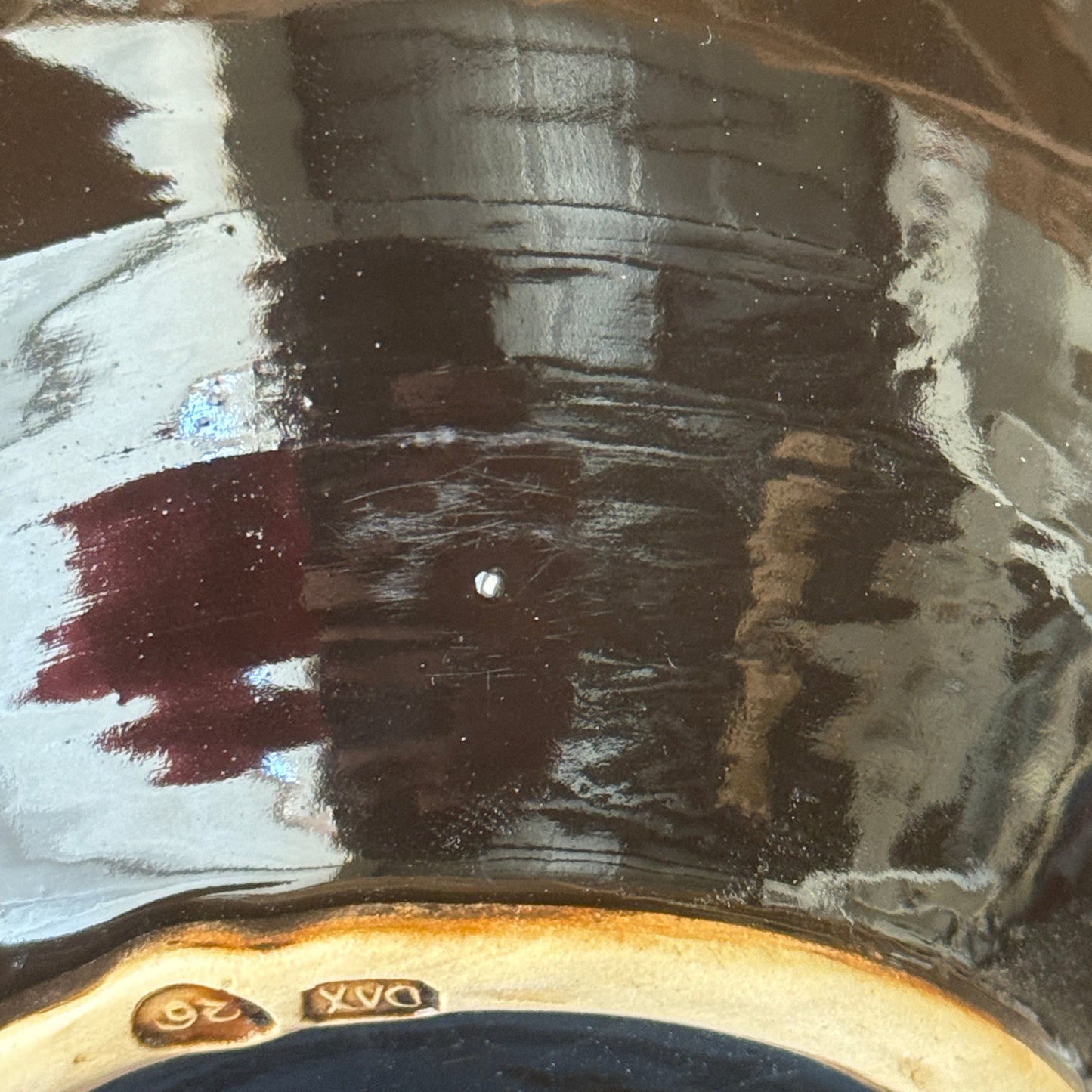 Close-up of a glossy dark brown ceramic surface with reflections, showing a small gold '26' mark and part of a painted design near the bottom edge.