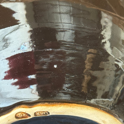 Close-up of a glossy dark brown ceramic surface with reflections, showing a small gold '26' mark and part of a painted design near the bottom edge.