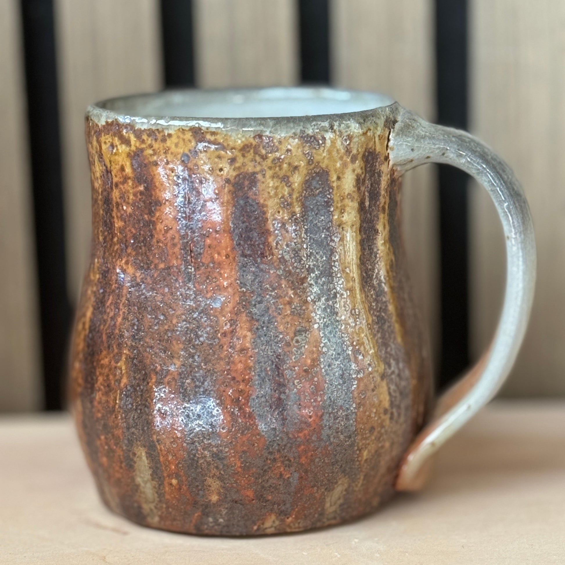 A rustic handmade ceramic mug with a textured, earthy glaze featuring brown, orange, and gray tones, set against a neutral wooden background.