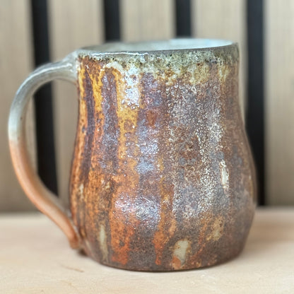 A rustic handmade ceramic mug with a textured, oxidized finish showing brown, orange, and silver tones, set against a neutral background.
