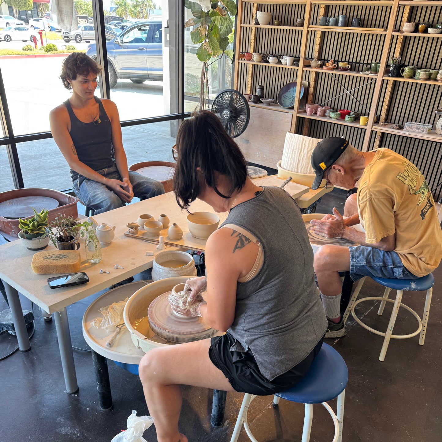 People working on pottery in a studio with shelves of finished ceramics.