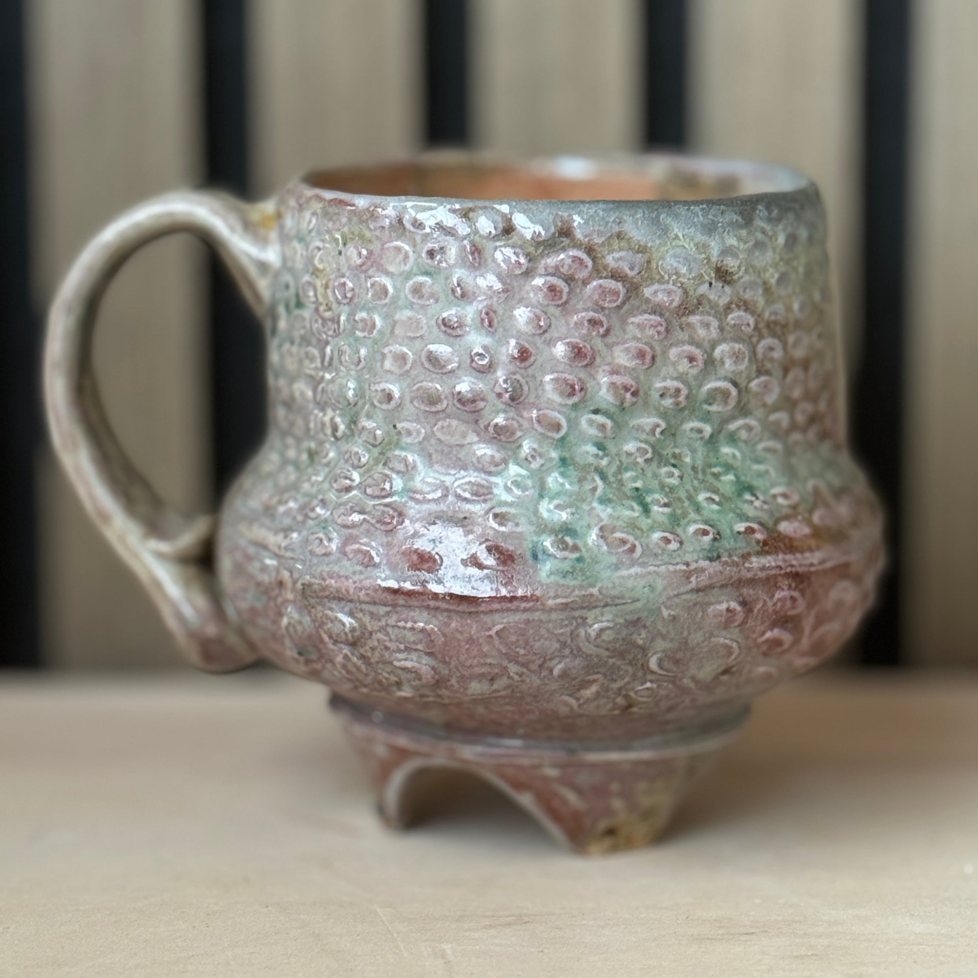 A handmade ceramic mug with a textured, hammered surface and a small footed base, featuring a glaze with pink, green, and brown tones, set on a light wooden surface.