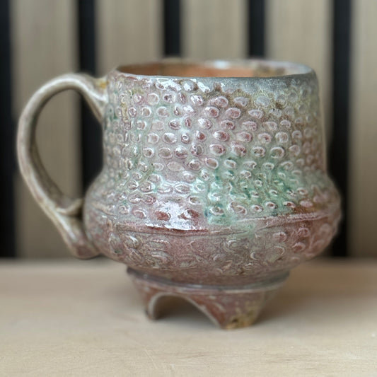 A handmade ceramic mug with a textured, hammered surface and a small footed base, featuring a glaze with pink, green, and brown tones, set on a light wooden surface.