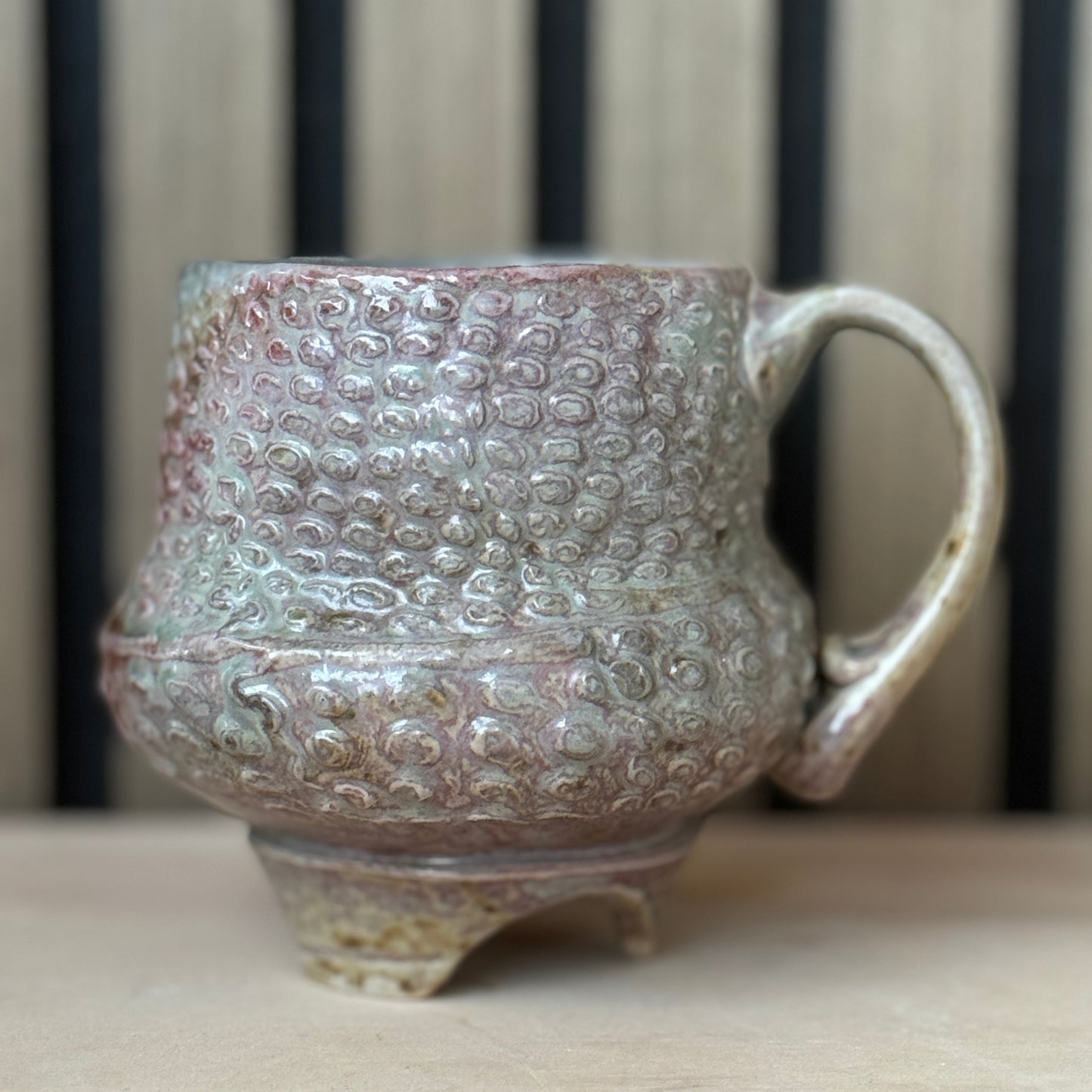 A handmade ceramic mug with a textured, bubble-like embossed pattern and a rustic footed base, featuring a pinkish-brown and sage green glaze, set against a blurred background of vertical wooden slats.