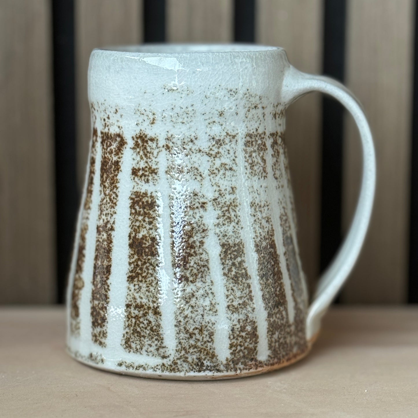 A handmade ceramic mug with a textured, ribbed design featuring vertical brown and white stripes, set against a neutral wooden background.