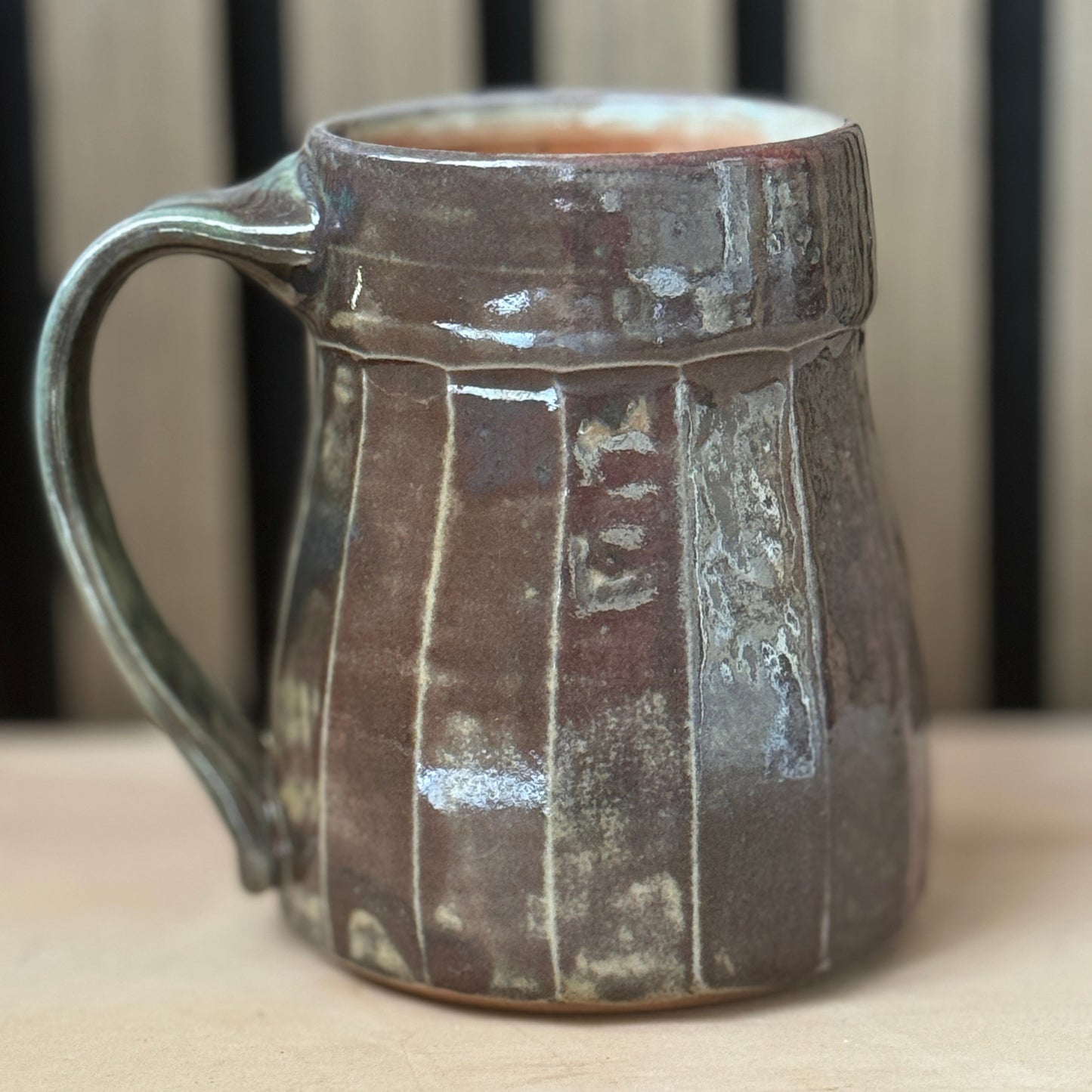 A handmade ceramic mug with a rustic, textured glaze in earthy brown and gray tones, featuring vertical ribbed details and a sturdy handle, set against a blurred background of vertical wooden slats.