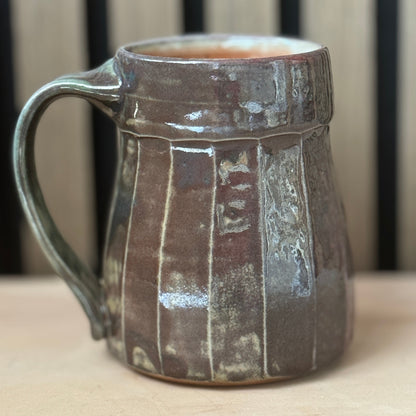 A handmade ceramic mug with a rustic, textured glaze in earthy brown and gray tones, featuring vertical ribbed details and a sturdy handle, set against a blurred background of vertical wooden slats.